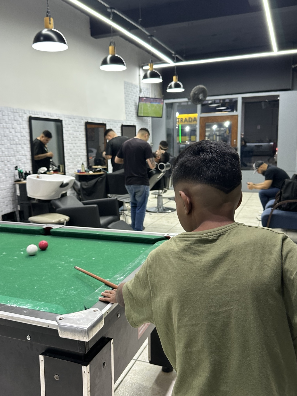 Cliente jogando sinuca dentro da Barbearia Calcki enquanto barbeiros atendem ao fundo